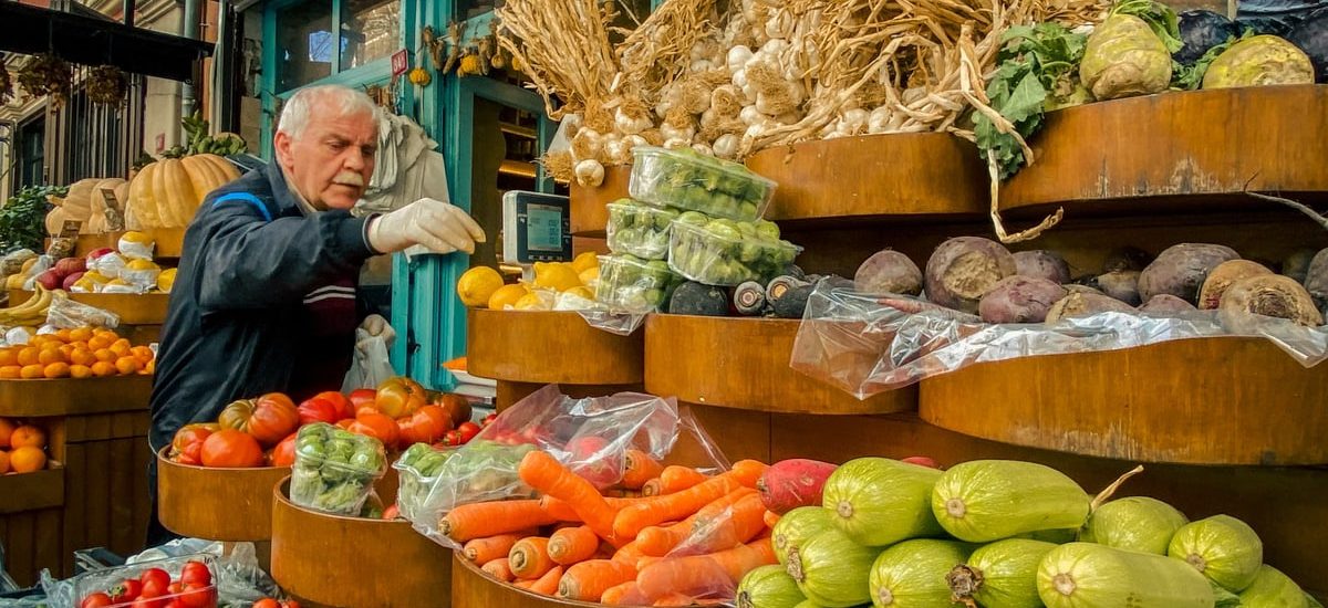 An elderly man selects vegetables at a vibrant outdoor urban market stand filled with fresh produce.