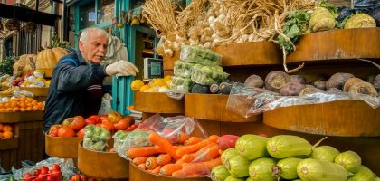 An elderly man selects vegetables at a vibrant outdoor urban market stand filled with fresh produce.