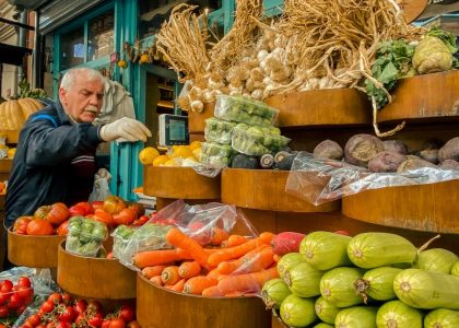 An elderly man selects vegetables at a vibrant outdoor urban market stand filled with fresh produce.