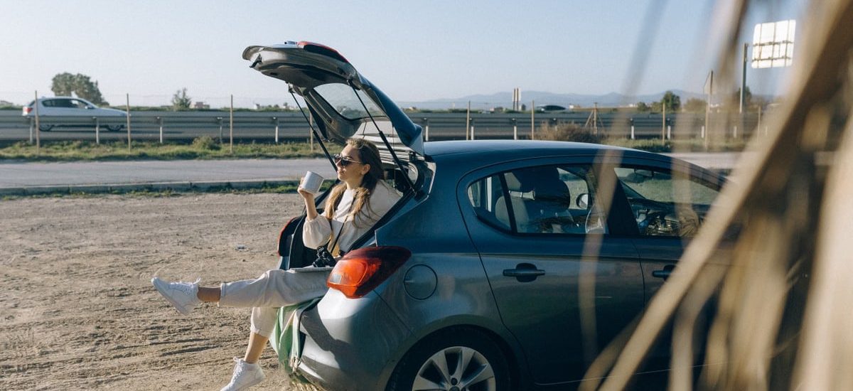 A woman sitting in the trunk of a parked car on a sunny day, enjoying a break during a road trip.