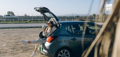 A woman sitting in the trunk of a parked car on a sunny day, enjoying a break during a road trip.
