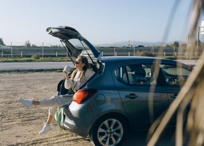 A woman sitting in the trunk of a parked car on a sunny day, enjoying a break during a road trip.